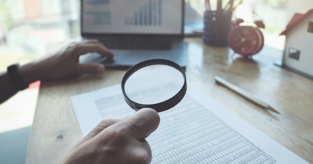 person holding a magnifying glass over documents, open laptop